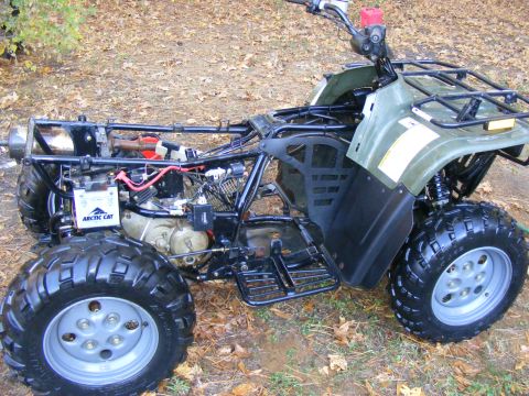 Green Arctic Cat ATV side view, engine and battery visible, parked on leaf-covered ground.
