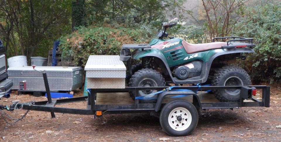 Green Polaris Xplorer 4x4 ATV strapped to a black utility trailer, side view, with diamond-plate toolbox