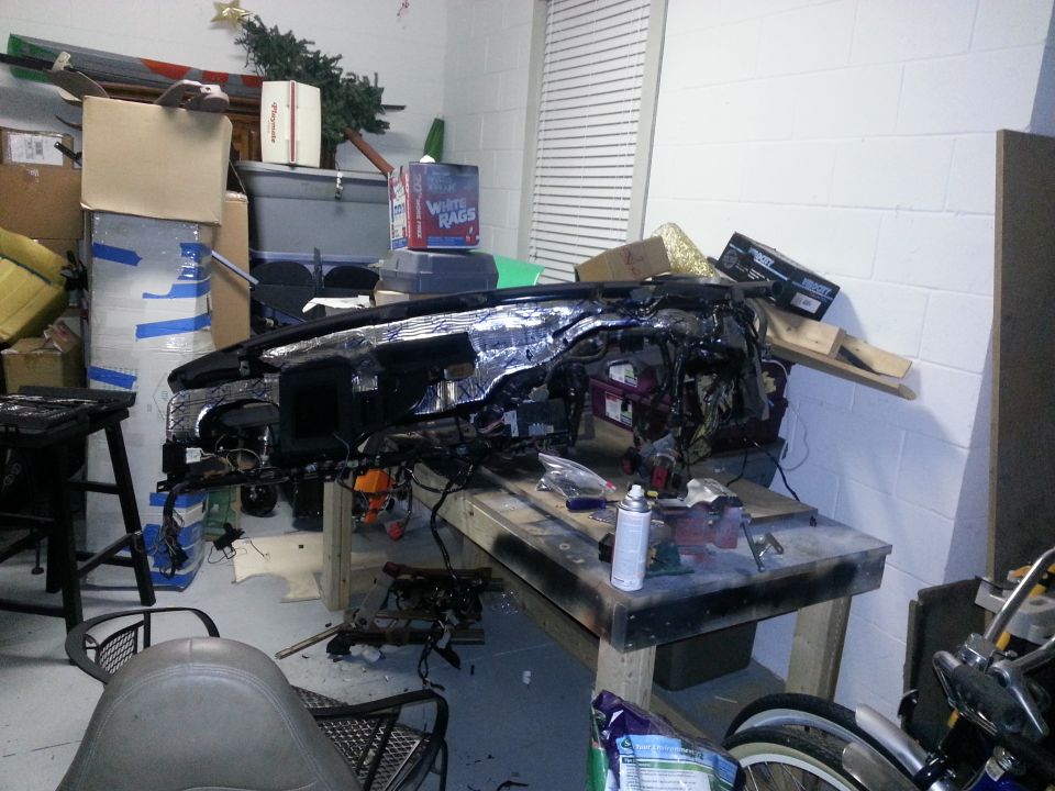 Disassembled vehicle dashboard with exposed wiring and insulation, resting on a workbench