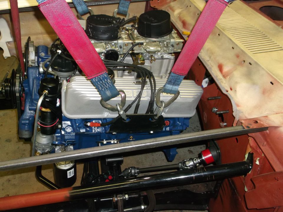 Blue V8 engine with dual carburetors and silver valve covers, secured by red lifting straps.