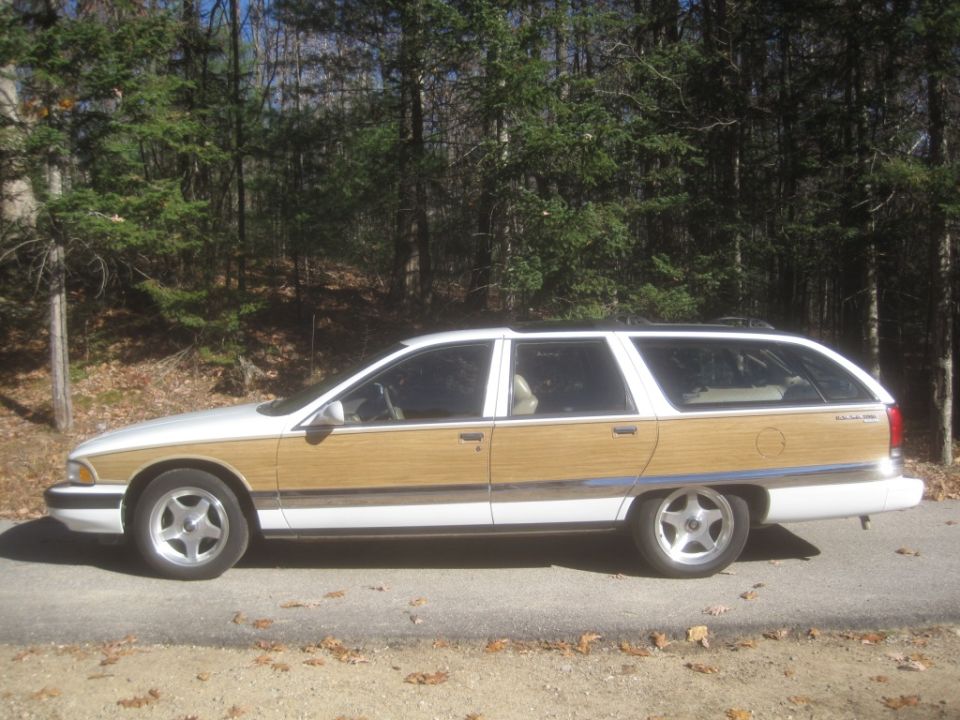 White and wood-paneled station wagon parked on a road with trees in the background.