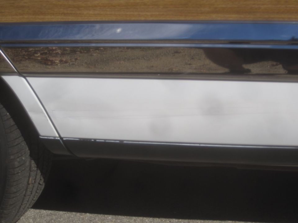 Close-up of a car's lower side panel, showing white paint with scratches and a black trim piece.