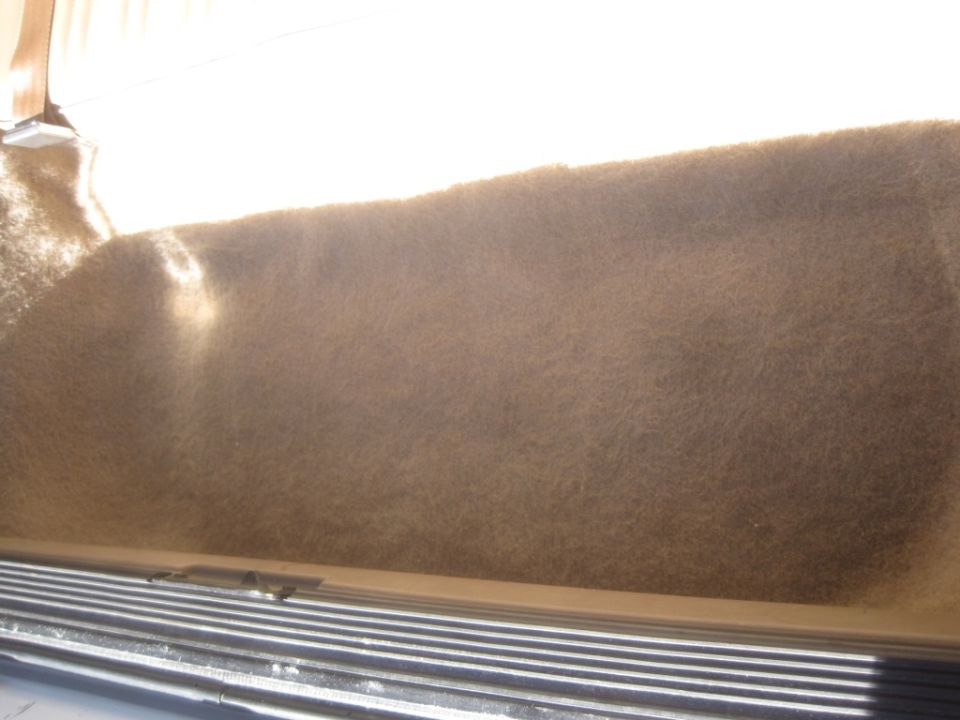 Interior of a vehicle with brown carpet and metal blinds, illuminated by sunlight.