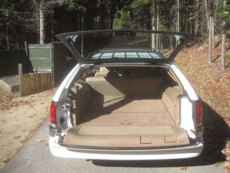 White station wagon with open rear hatch, showing tan interior, parked in a wooded area.