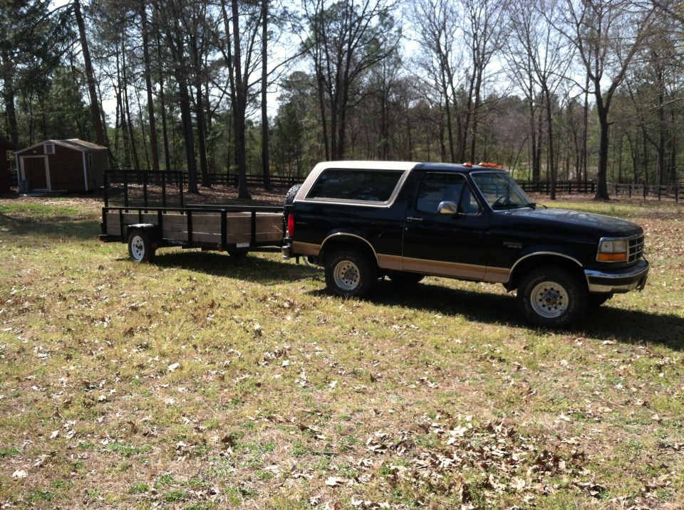Black Ford Bronco with tan top and trailer in a grassy yard with trees and a shed.