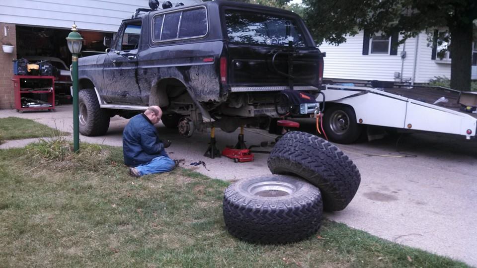 Man working on a black Ford Bronco in a driveway with tires and a car hauler trailer.