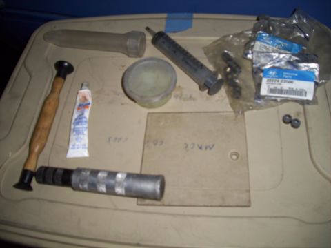1995 Ford Probe - A collection of tools and parts are laid out on a beige surface. Visible items include a syringe, a tube of sealant, a hammer-like tool, an…