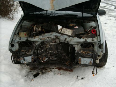 1986 Ford Taurus - The front end of a 1986 Ford Taurus is heavily damaged, revealing the engine compartment with the hood open.