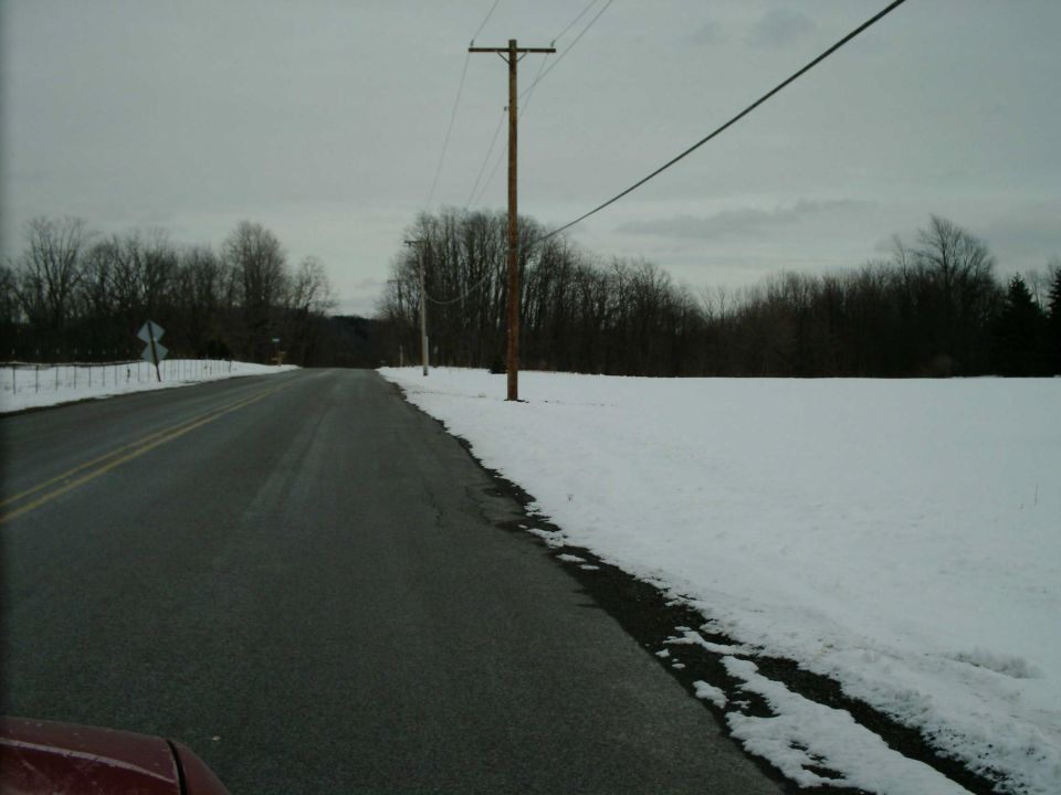 Two-lane rural road with snow-covered fields and utility poles; edge of a dark red hood visible at bottom left