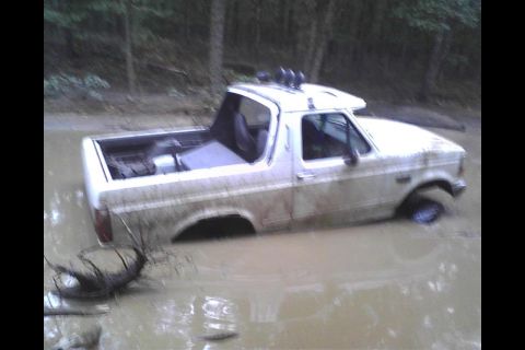 1994 Ford Bronco - This white SUV is partially submerged in a muddy body of water, with trees visible in the background.