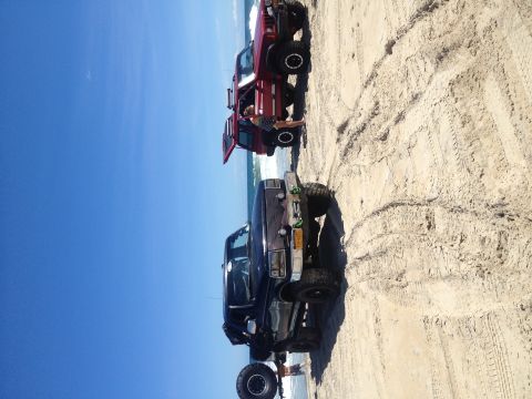 1995 Ford Bronco - Two off-road vehicles, a Ford F-Series truck and a Jeep Wrangler, are parked on a sandy beach with the ocean in the background.
