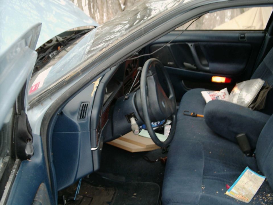 Driver's side interior of a light blue Ford sedan with blue cloth seats, open door, debris and open hood visible