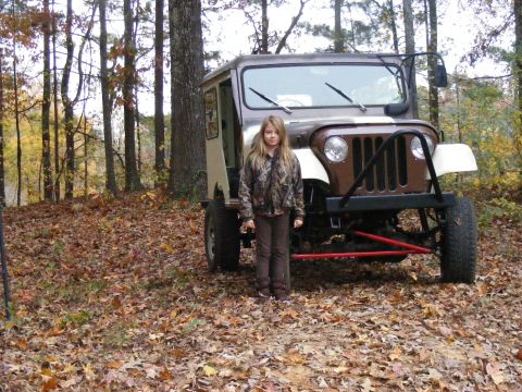 Girl in camouflage standing next to a custom brown and tan off-road vehicle in a forest.