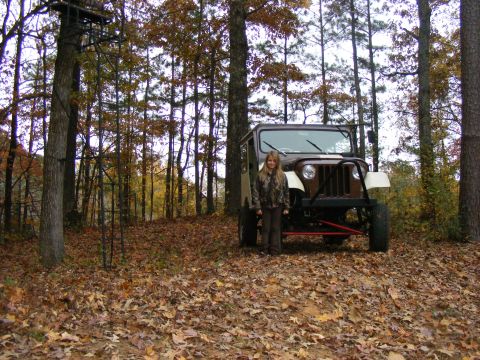 Young woman in camouflage standing by a brown and white Jeep in a forest.