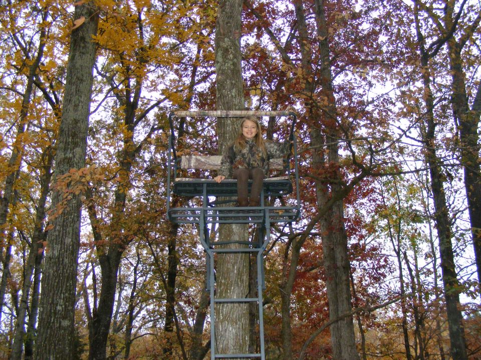 A young girl in camouflage sits in a deer stand among trees with fall foliage.