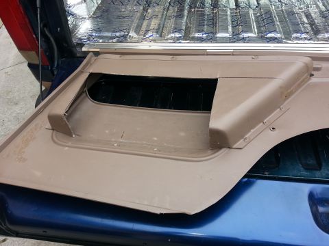 1994 Ford Bronco - A tan interior panel with a large cutout is shown, possibly for a speaker or storage, vent, in the bed of a blue truck.