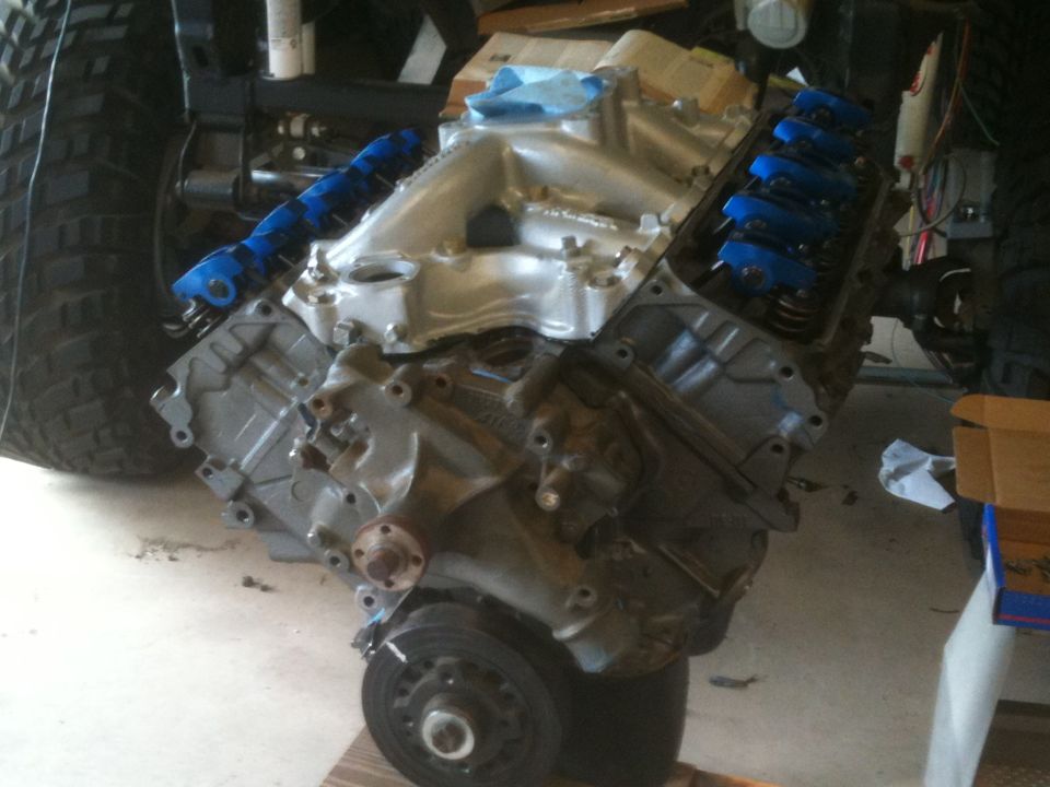 V8 engine block with silver intake manifold and blue valve covers, next to a large tire.