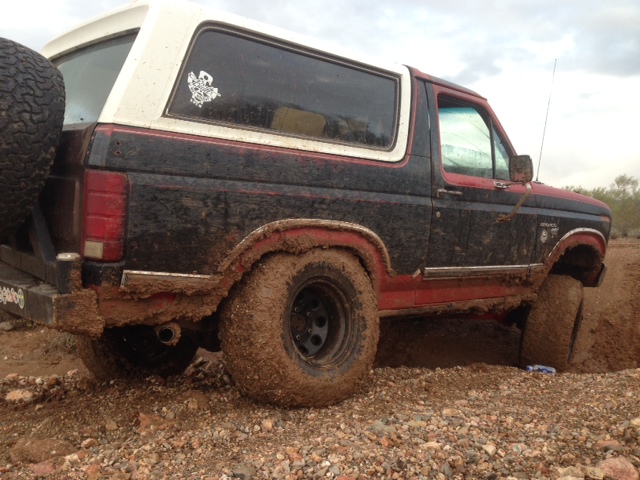 Mud-covered black and red Ford Bronco with a white top, stuck in dirt