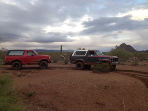 1996 Ford Bronco - Two Ford Broncos, one red and one two-tone, are parked side-by-side on a dirt road in a desert environment under a cloudy sky.