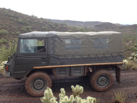 1996 Ford Bronco - Military truck with canvas top navigates a desert trail with cacti and hills in the background.