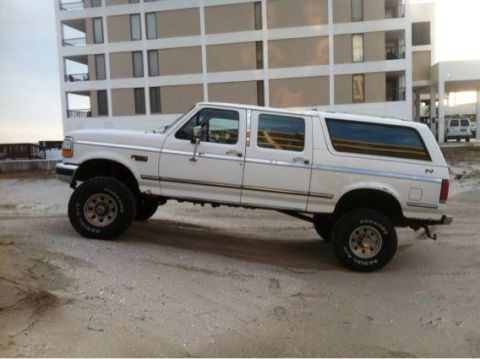 1995 Ford Bronco - This white 1995 Ford Bronco extended cab pickup truck with a camper shell is parked on a sandy surface near a building.