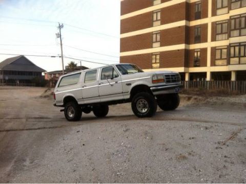 1995 Ford Bronco - This lifted SUV is parked on a sandy lot with a large brick building and some houses visible.