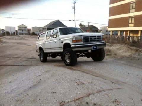 1995 Ford Bronco - A lifted white Ford Bronco SUV is parked on a sandy road near beach houses and a tall building.