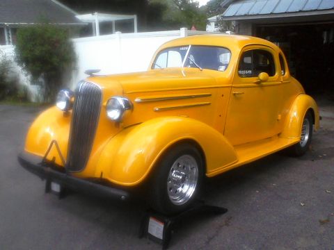 1935 Chevrolet New - A bright yellow 1935 Chevrolet coupe is shown from a three-quarter front view, parked in front of a garage.