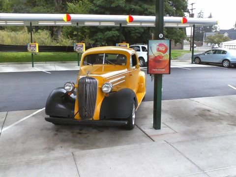 1935 Chevrolet New - A yellow 1935 Chevrolet New is parked at a drive-thru, with a white SUV and a blue sedan visible in the background.