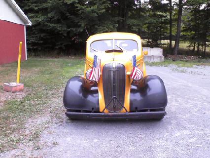 1935 Chevrolet New - A yellow truck with American flags is parked in front of a red barn and trees.