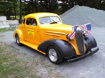 1935 Chevrolet New - This yellow coupe, likely a 1936 Chevrolet, is adorned with American flags on its distinctive grille.