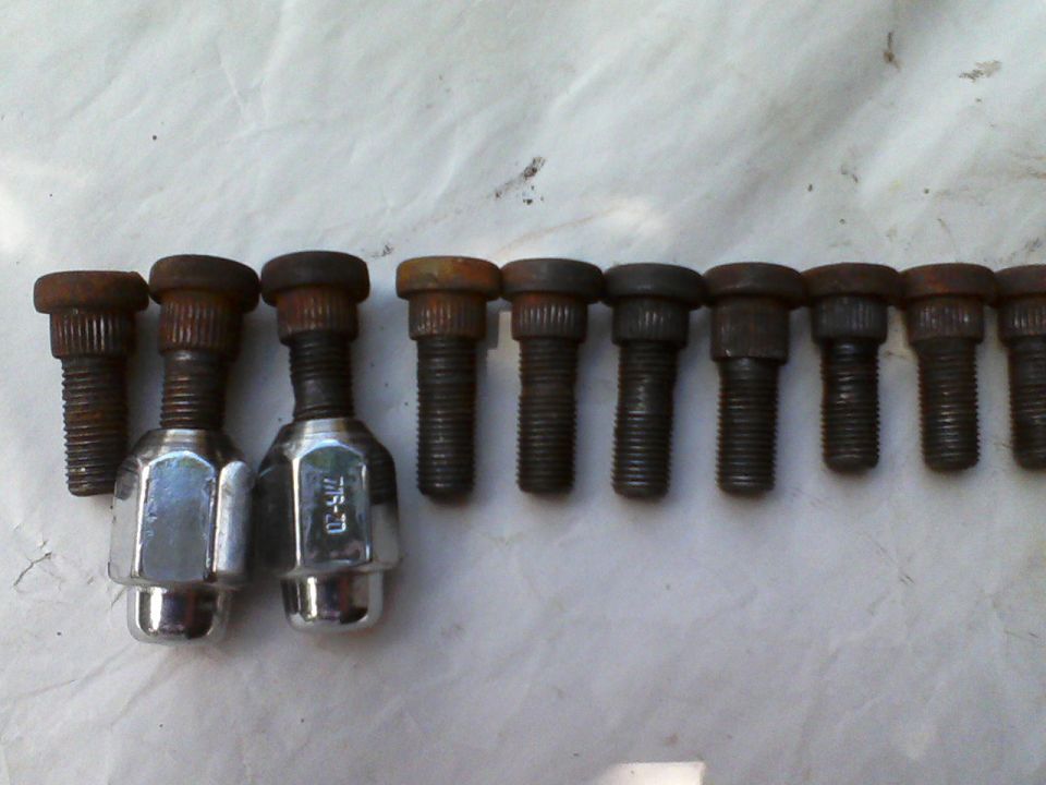 Lug nuts and bolts on a white surface. Two chrome lug nuts are on the left, marked 7/16-20. Eight rusty bolts are to their right