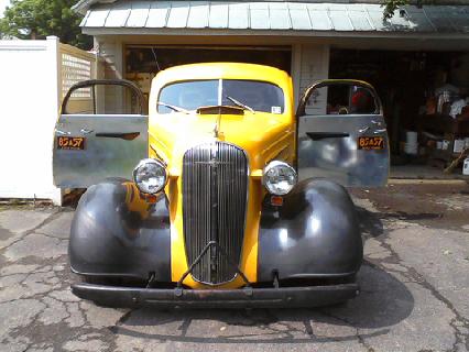 1935 Chevrolet New - A yellow truck with its doors open sits in front of a garage, displaying a license plate.