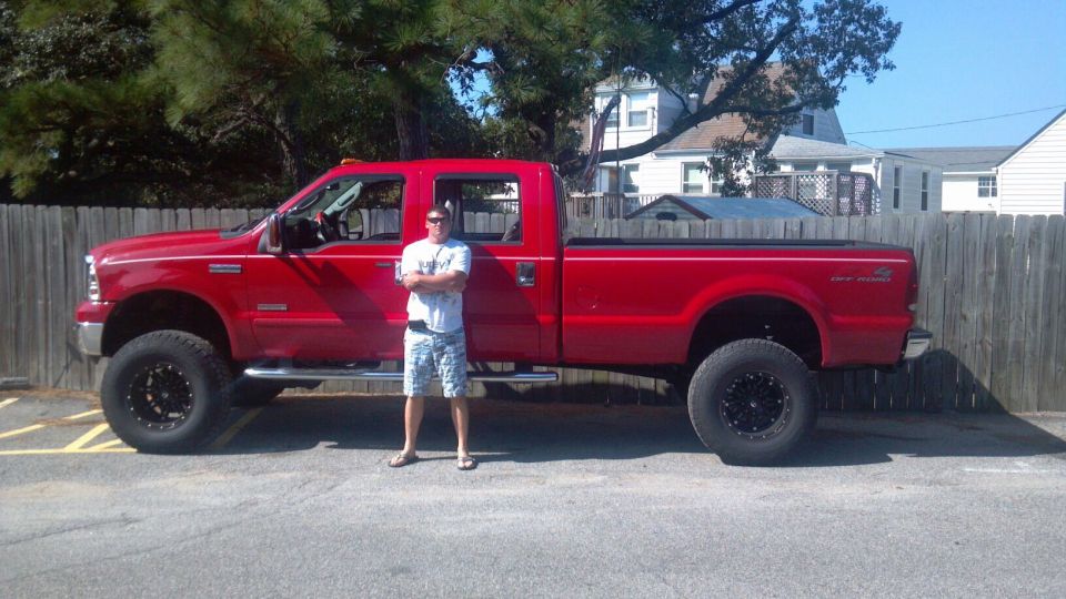 Red 2007 Ford F-350 Super Duty pickup truck, lifted with large black wheels, parked next to a wooden fence
