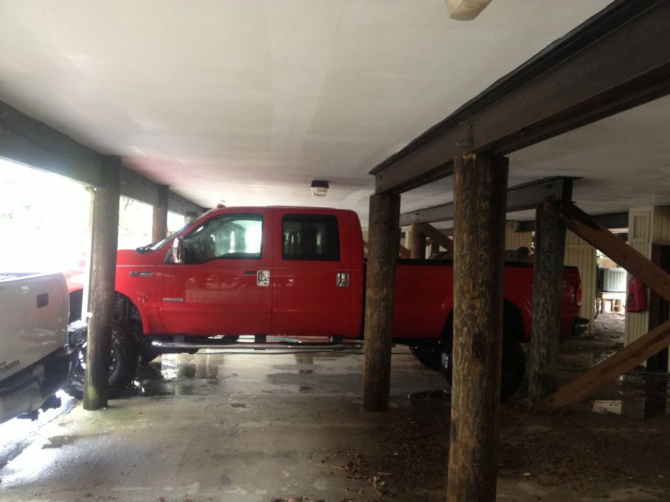 Red crew cab pickup truck parked under a structure with wooden posts. Another truck is partially visible on the left