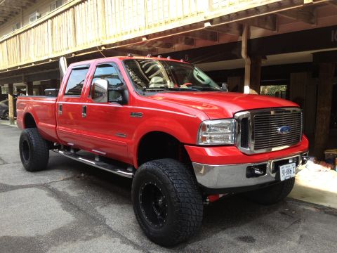 2007 Ford F-350 - This red pickup truck is parked outside a building with wooden siding. It has large off-road tires and a prominent exhaust pipe.