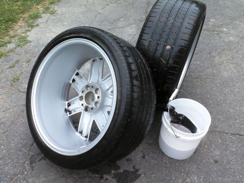 2004 Chrysler Crossfire - Two car wheels and tires are laid out next to a bucket of water on a paved surface.