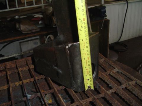 1993 Ford Bronco - A dark metal radius arm bracket is shown with a yellow measuring tape for scale. The part appears to be for a vehicle's suspension system.