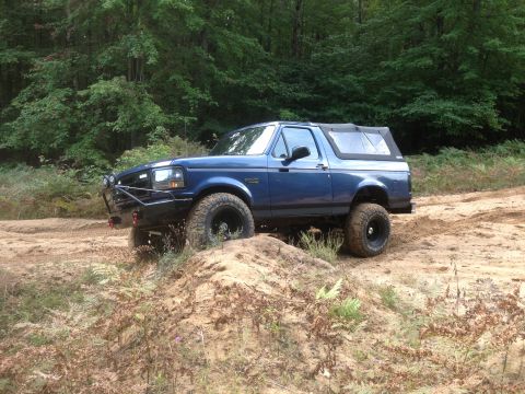 1996 Ford Bronco - This blue Ford Bronco is navigating a dirt path with dense trees in the background.