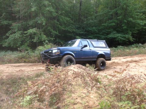1996 Ford Bronco - A blue 1996 Ford Bronco navigates a dirt trail with dense woods in the background.