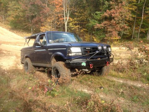 1996 Ford Bronco - This blue 1996 Ford Bronco is equipped with a custom front bumper and off-road tires, navigating a dirt path surrounded by autumn foliage.