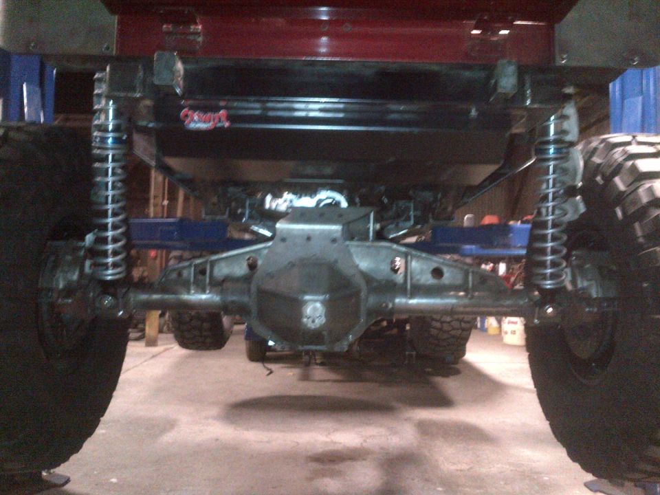 Rear axle assembly with large coilover shocks and massive tires. A red body panel is visible above