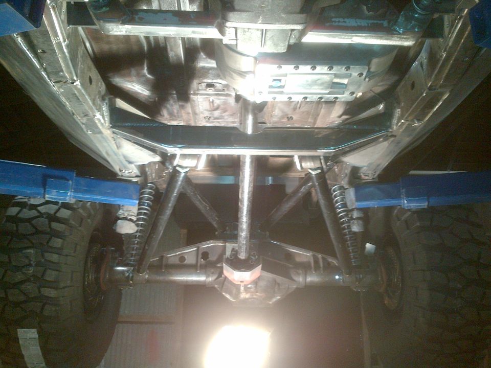Underside of a vehicle on a lift, showing suspension and drivetrain components, with large off-road tires