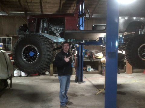 1985 Jeep CJ7 - A heavily modified Jeep is suspended on a lift in a workshop, with a man standing next to it.