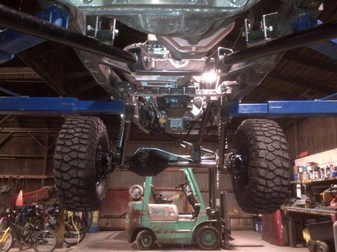1985 Jeep CJ7 - The underside of a Jeep is viewed from below while on a lift, with a forklift positioned underneath.