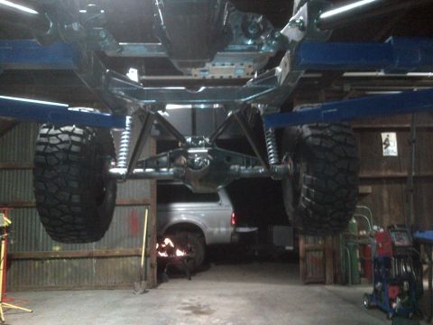 1985 Jeep CJ7 - The underside of a vehicle is shown on a lift, revealing its suspension and large off-road tires, with another vehicle partially visible in…