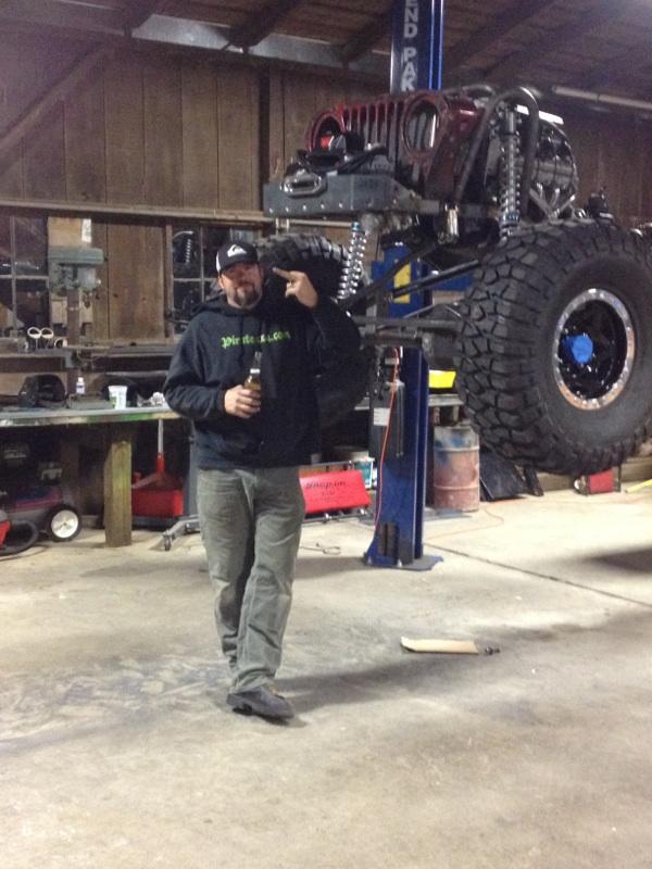 Man in garage points at heavily modified Jeep lifted in the air. It has large tires and custom suspension