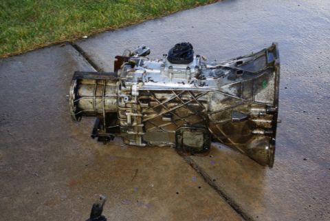 1995 Ford Bronco - A used transmission assembly rests on a wet concrete surface next to a patch of grass.