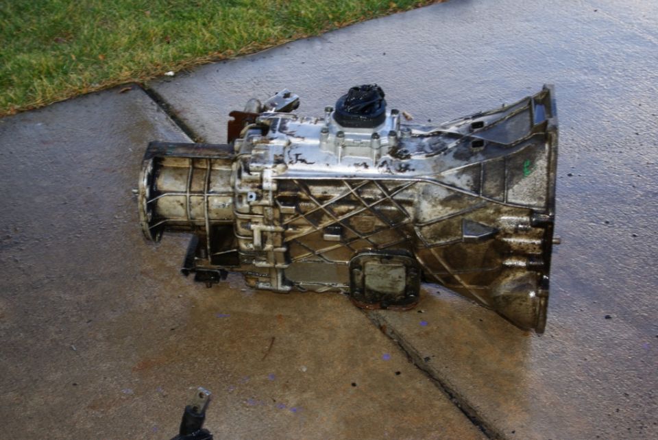 Dirty transmission assembly on wet concrete, with grass in background
