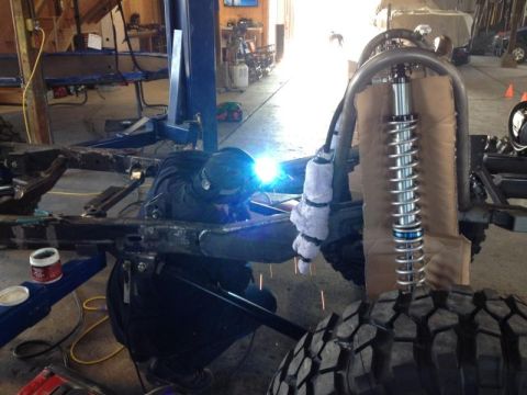 1985 Jeep CJ7 - A welder works on the frame of a vehicle, with suspension and tire components in view.
